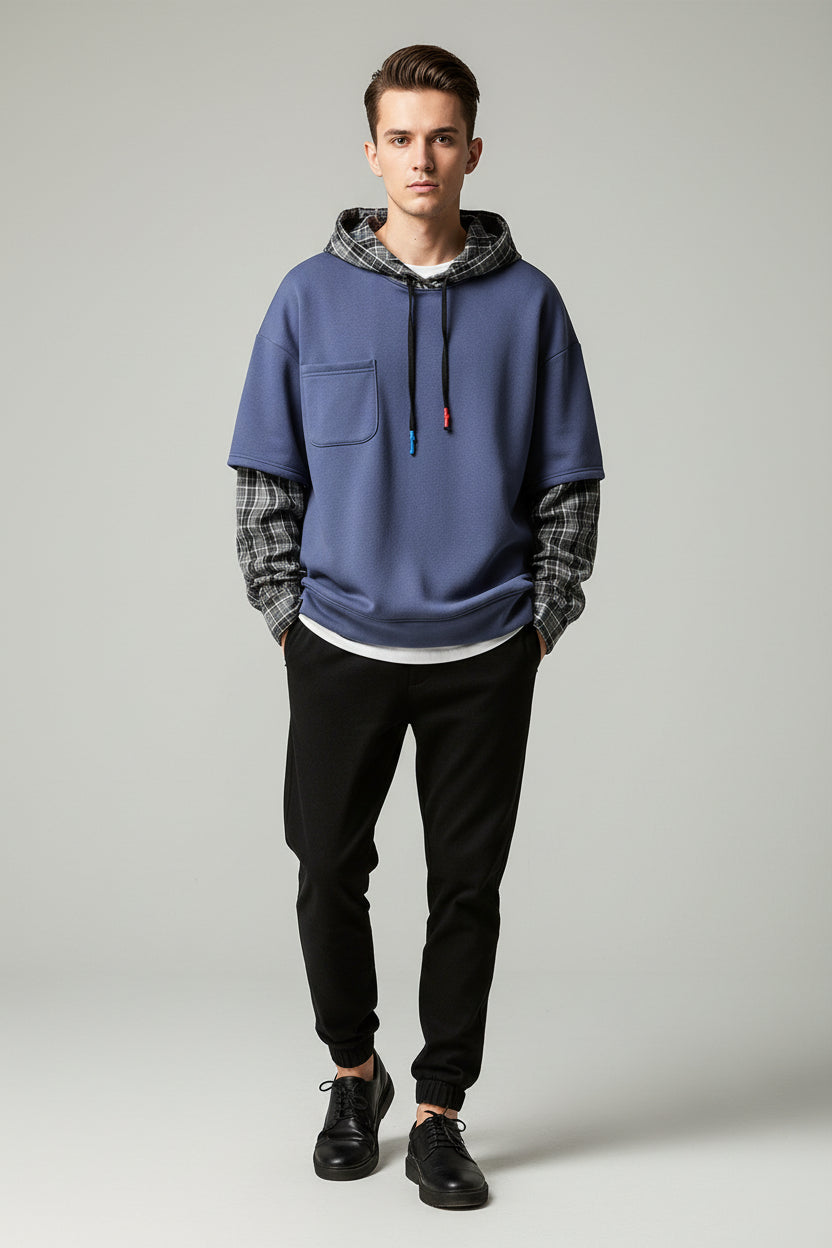 A blue pullover hoodie with a plaid hood lining and matching plaid rolled sleeves, hanging on a black hanger against a faux stone wall background.