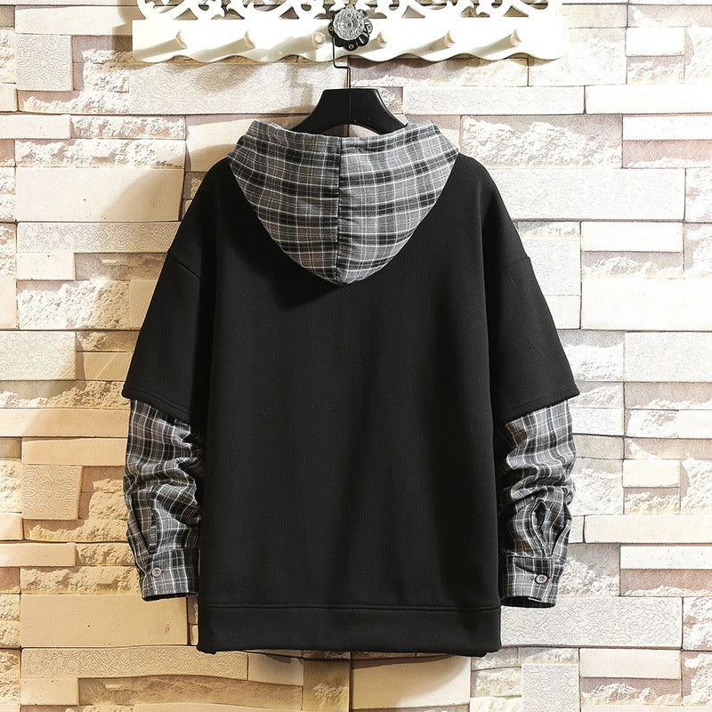 A black pullover hoodie with a gray and white plaid hood lining and matching plaid cuffed sleeves, hanging on a black hanger against a light beige faux brick wall.