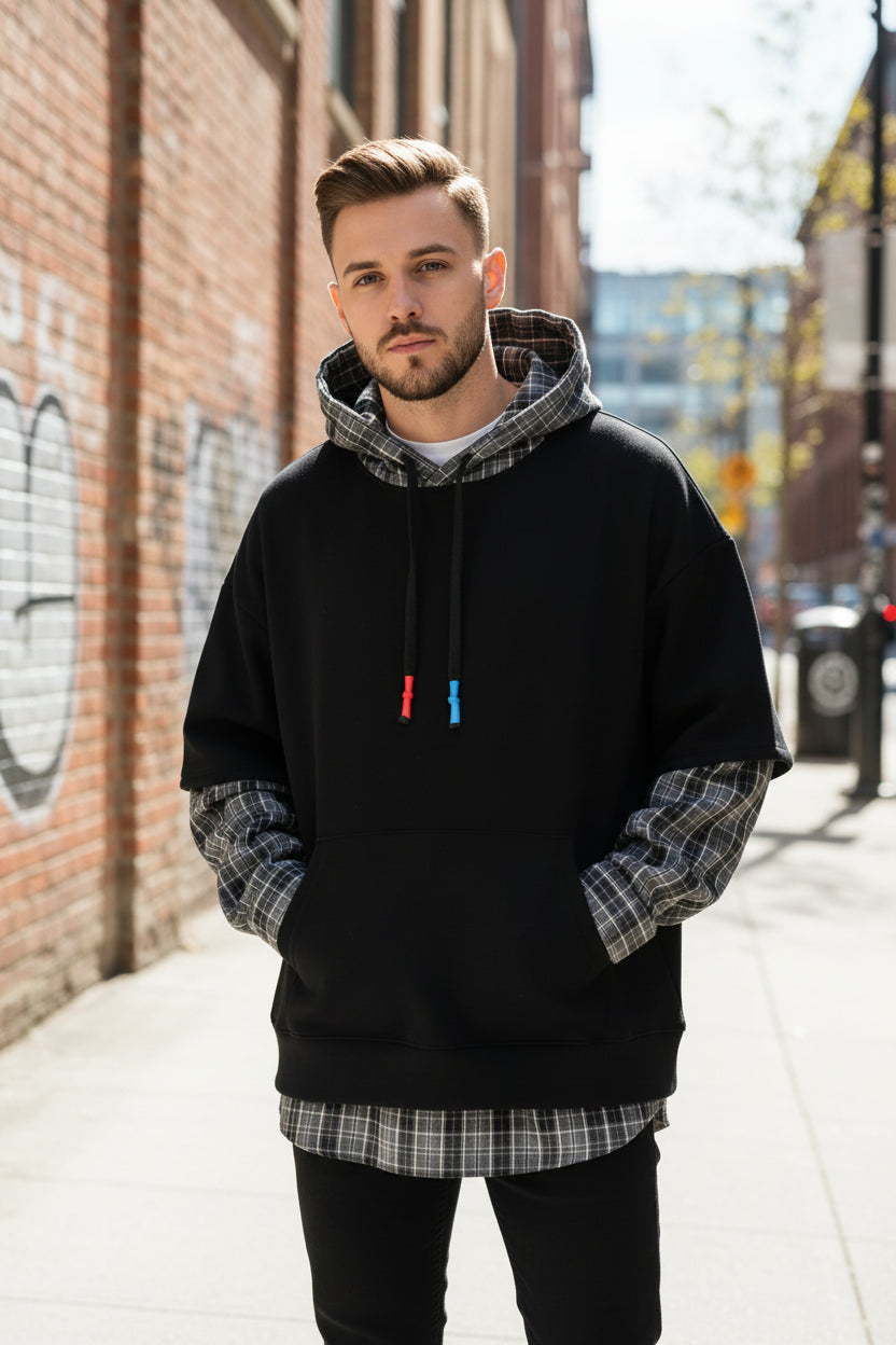 A black pullover hoodie with a plaid hood lining and matching plaid sleeves, featuring a small red embroidered detail on the chest and a blue drawstring tip, displayed against a dark wood-paneled background.