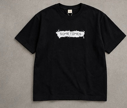 A black short-sleeve crewneck t-shirt with a white distressed graphic on the chest reading 'SOMETIMES.' in a paint-splatter style, laid flat on a textured gray background.