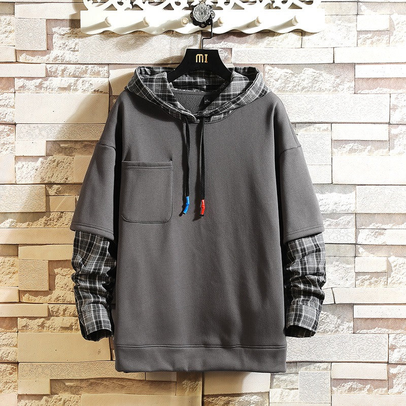 A dark gray pullover hoodie with a plaid patterned hood and matching plaid cuffed sleeves, hanging on a black hanger against a light-colored brick wall.